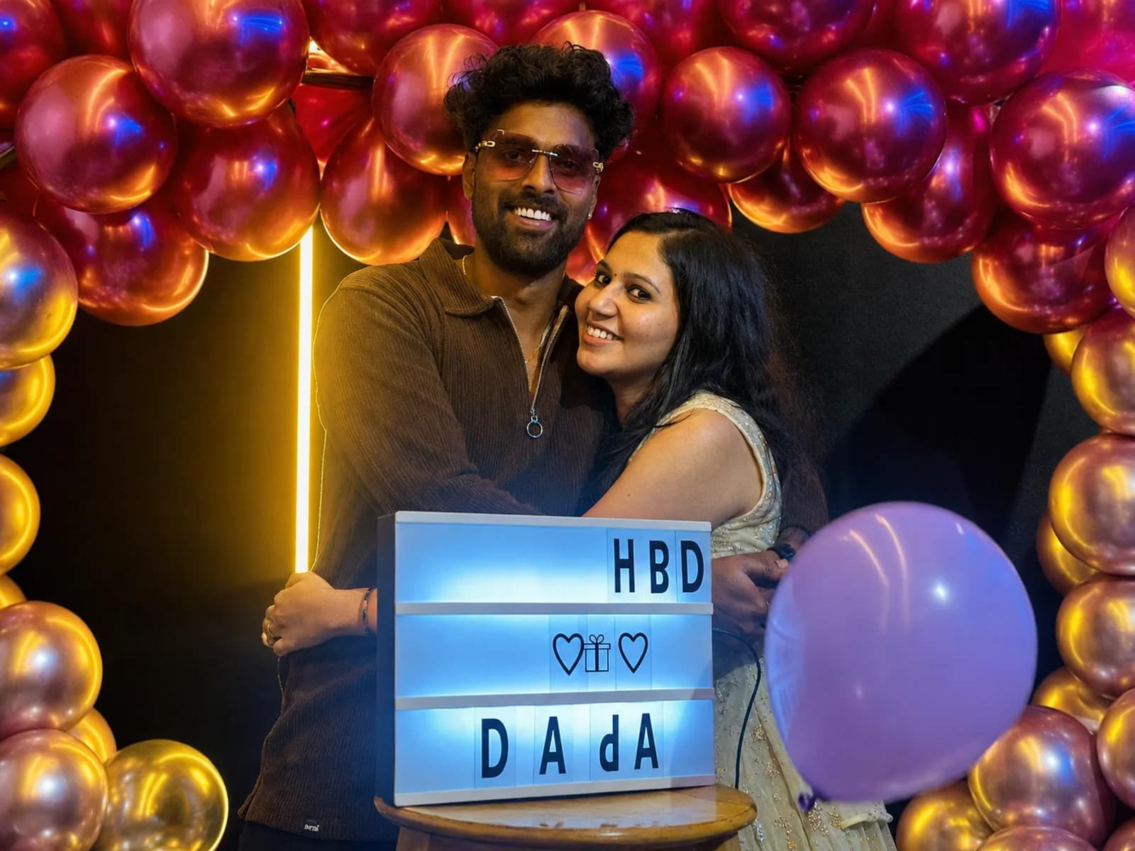 A couple embracing beside a lit HBD DADA letter board, under a red and gold chrome balloon arch