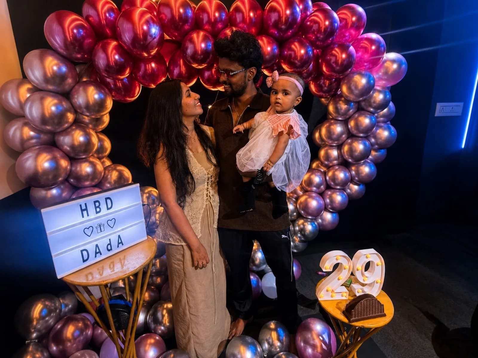 A family with a small child under a pink and chrome heart balloon arch, with a lit letter board, a lit 29 and a cake