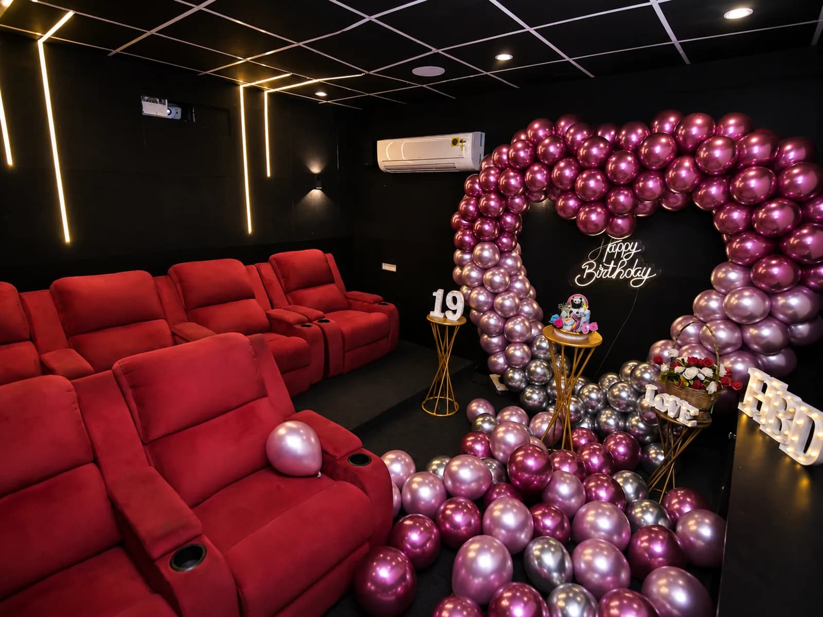 Rows of red recliner seats facing a pink and chrome heart balloon arch with a neon Happy Birthday sign, a cake stand and lit LOVE and HBD letters