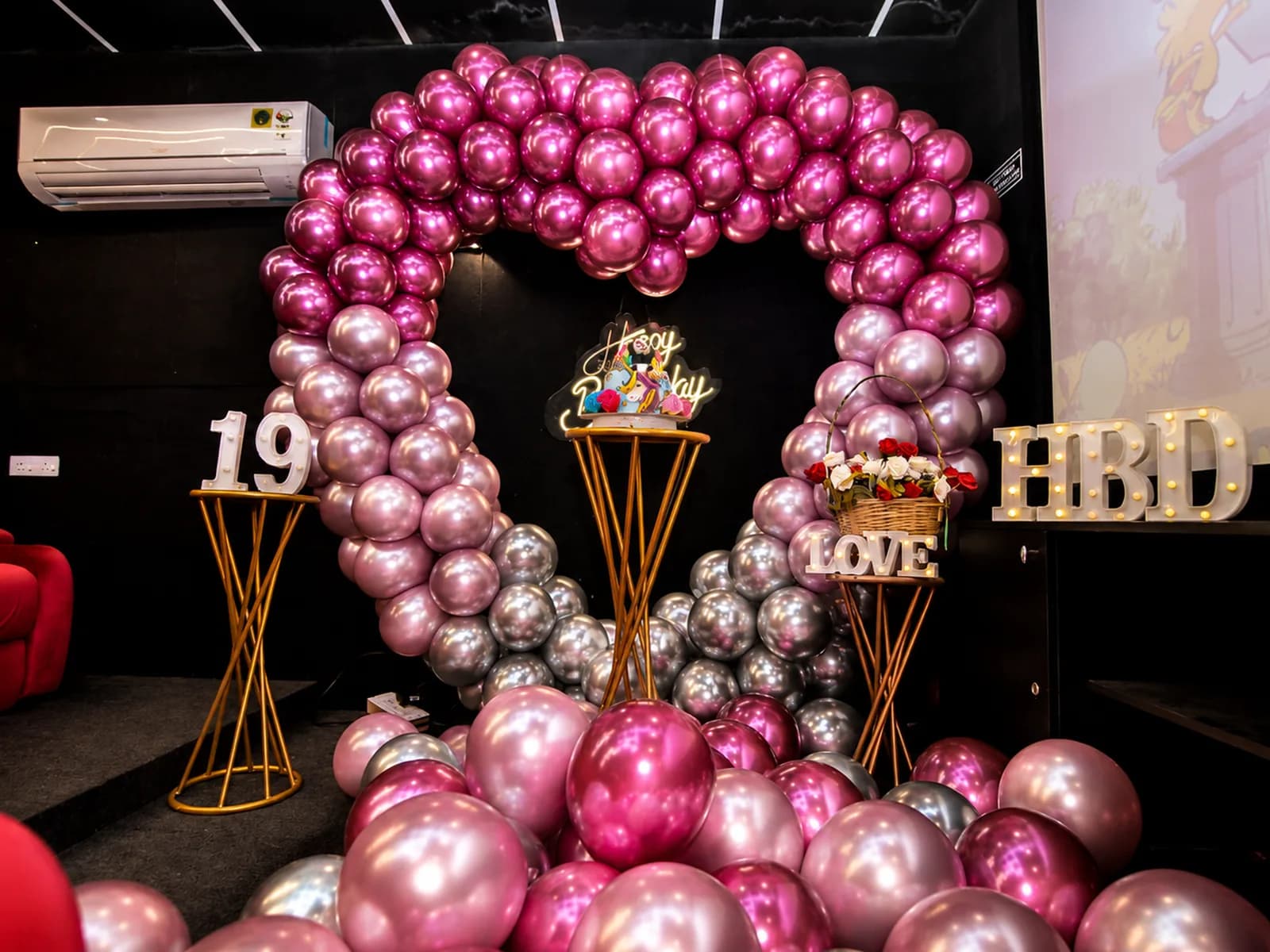 A pink and chrome heart balloon arch around a neon Happy Birthday sign, with a cake stand, a rose basket and lit 19, LOVE and HBD letters