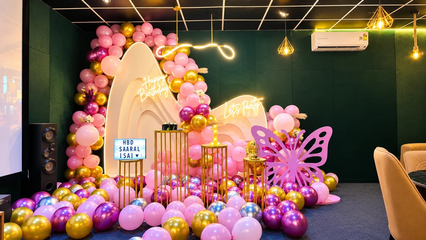 A pink and gold balloon arch around a cream sculpted backdrop with neon Happy Birthday and Let's Party signs, a pink butterfly cutout and a lit 1 sign, against a dark green velvet wall