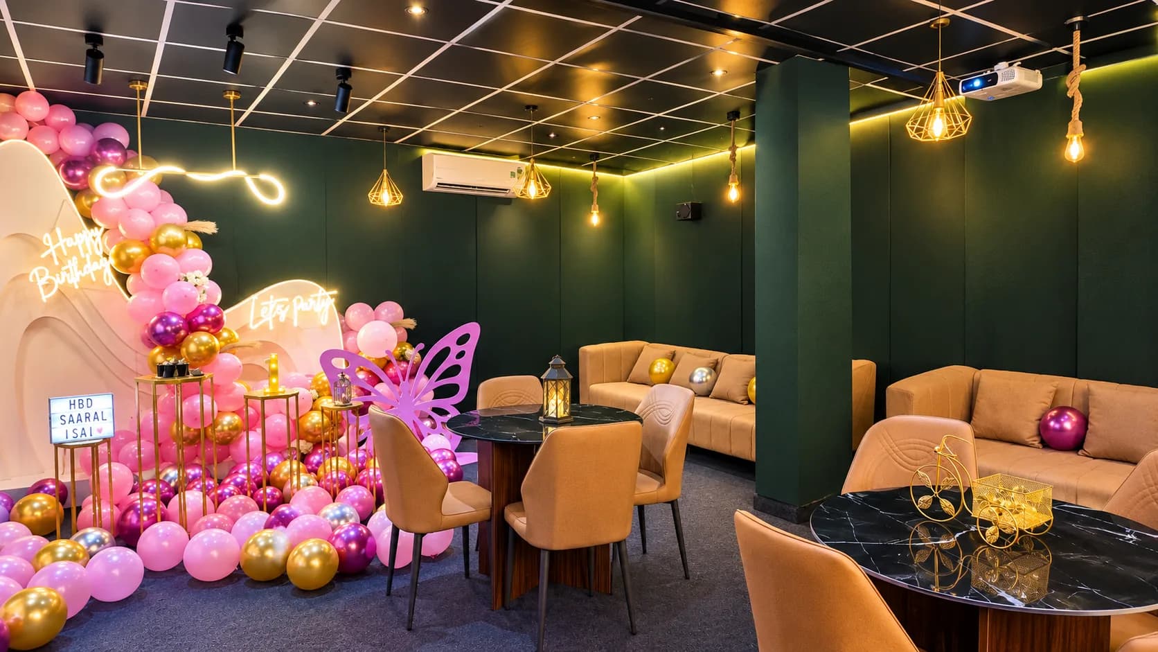 Tan sofas and chairs around black marble tables in a dark green velvet room with cage pendant lamps, beside a pink and gold balloon backdrop with a pink butterfly cutout
