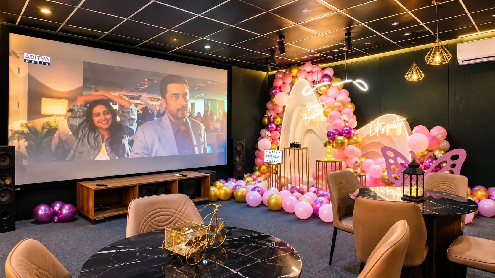 The room seen from the corner: the projection screen and floor speakers on the left, tan chairs around black marble tables, and a pink and gold balloon backdrop on the dark green wall