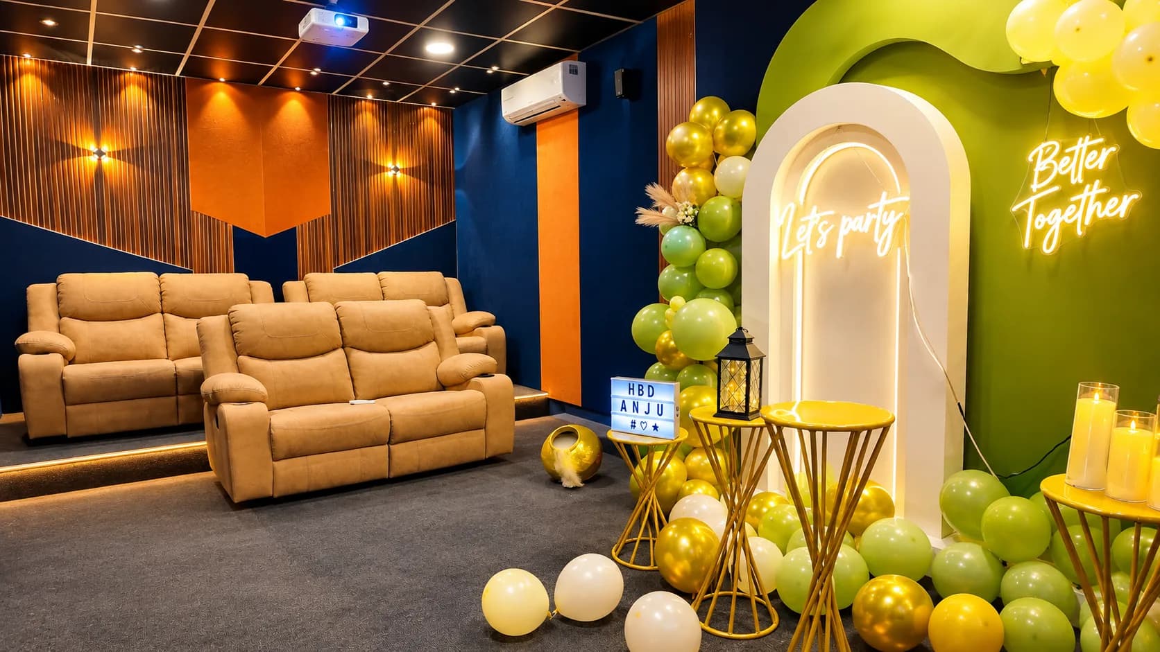 Three tan recliner sofas on tiered platforms in a navy and orange room with wood panelling, facing a green and gold balloon arch and a neon Let's Party sign