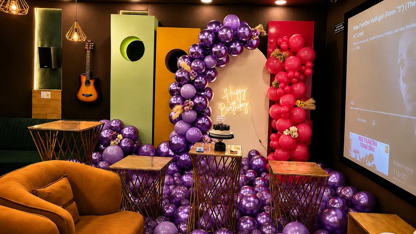 A purple and magenta balloon arch around a cream backdrop with a neon Happy Birthday sign and a cake, beside a red balloon column, an acoustic guitar on the wall and a tan armchair