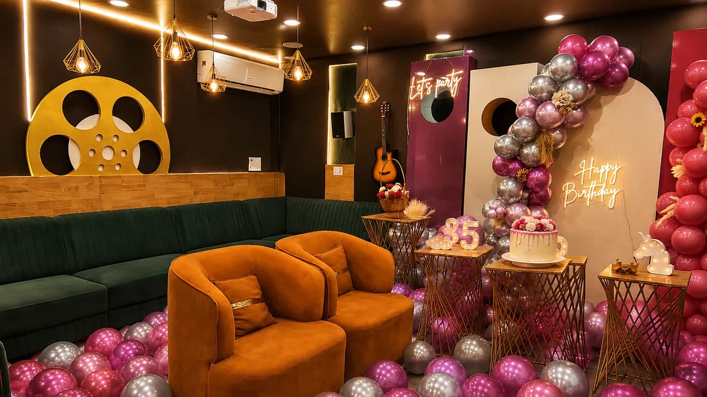 The whole room seen from the door: a green corner sofa and two tan armchairs standing in pink and silver floor balloons, under pendant lights and a gold film-reel wall sign