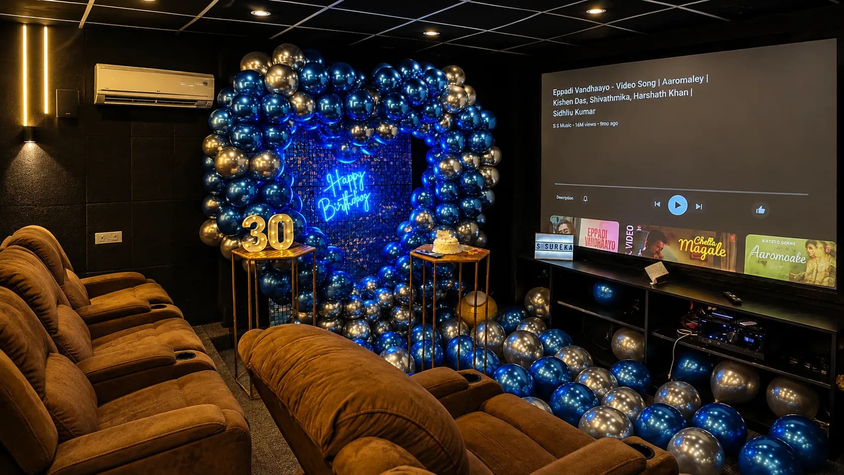 A blue and silver chrome balloon arch over a sequin wall with a neon Happy Birthday sign, a lit 30 sign and a cake on gold stands, beside the projection screen playing music videos