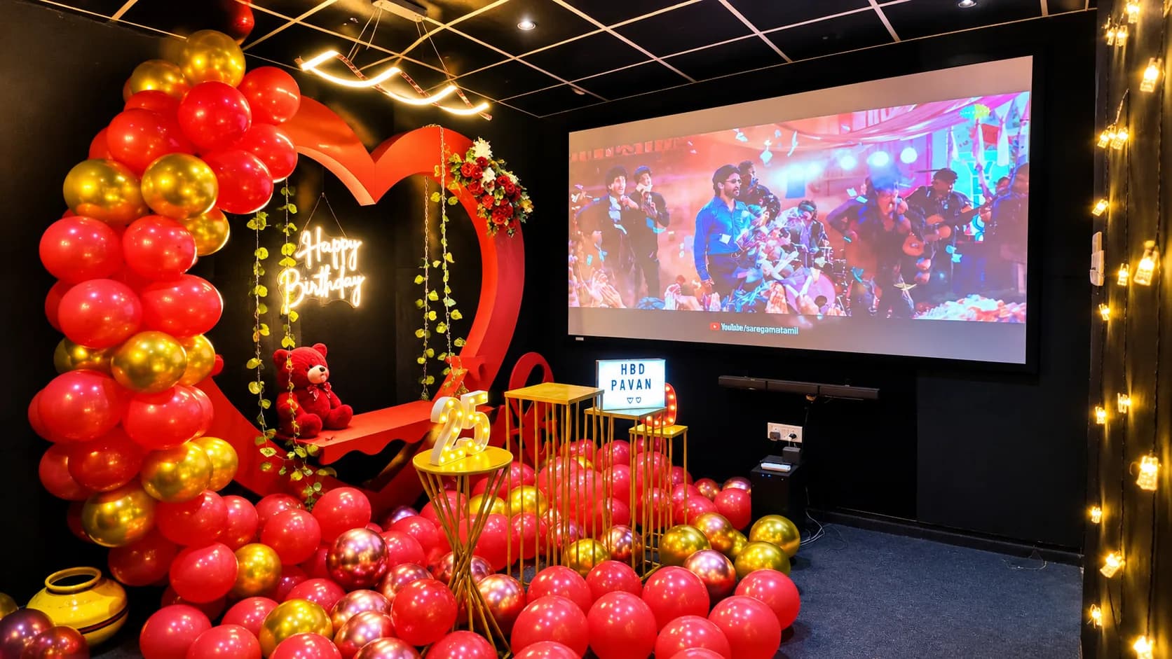 The projection screen playing a music video beside a red and gold balloon arch, a red heart backdrop with a gold swing and a teddy bear, and floor balloons across the carpet